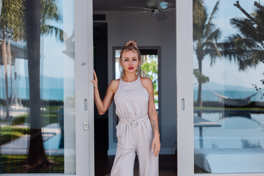 A stylish woman in a white jumpsuit stands poised before a door, exuding elegance and allure in an upscale setting.