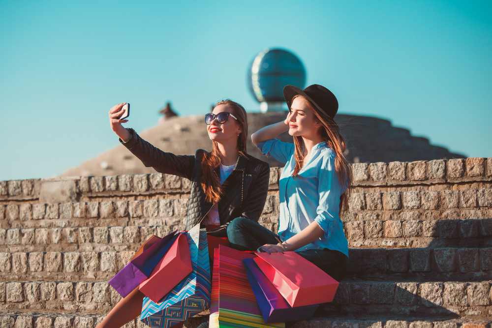 Two women sitting on a stone wall, holding shopping bags, while planning their dream vacation with Morocco travel packages.