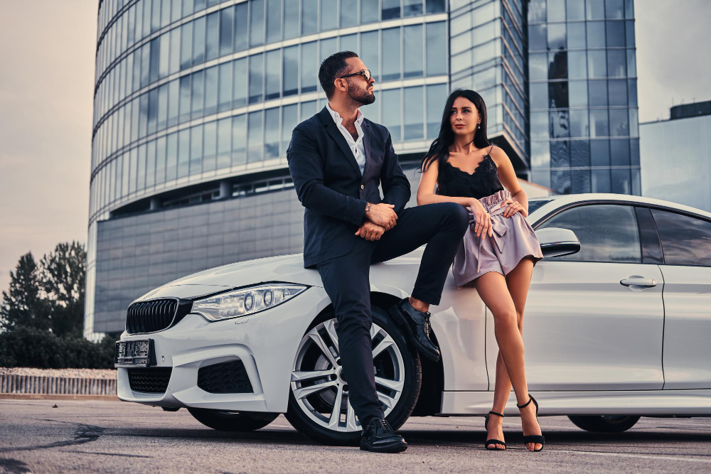 A couple in formal wear poses on a white car, embodying sophistication and style in Los Angeles.